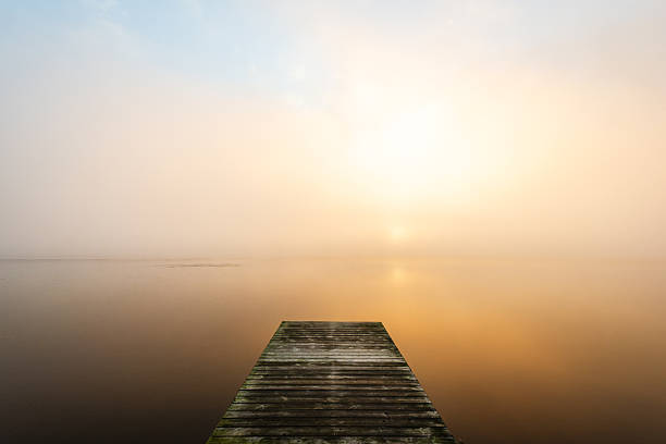 A long jetty stretches into a calm body of water, enveloped in soft mist under a golden sunrise. The peaceful setting invites reflection and serenity in Sweden's beautiful nature.