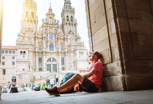 istock Joyful middle-aged backpacker pilgrim sitting on Obradoiro square in Santiago de Compostela, Spain, with Santiago de Compostela Cathedral â famous Camino de Santiago finish point. 2219087084