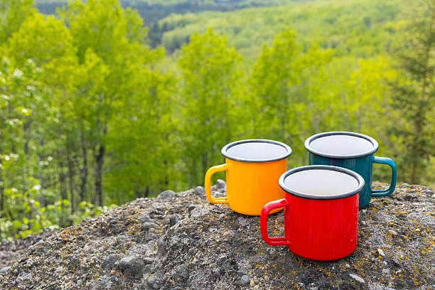 Three metallic tea cups-red, blue, and yellow-on ancient mountain rocks , set against a breathtaking view of a forested valley in sun light. Camping and hiking vibes of adventure.