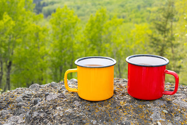Two metallic tea cups-red, and yellow-on ancient mountain rocks , set against a breathtaking view of a forested valley in sun light. Camping and hiking vibes of adventure.