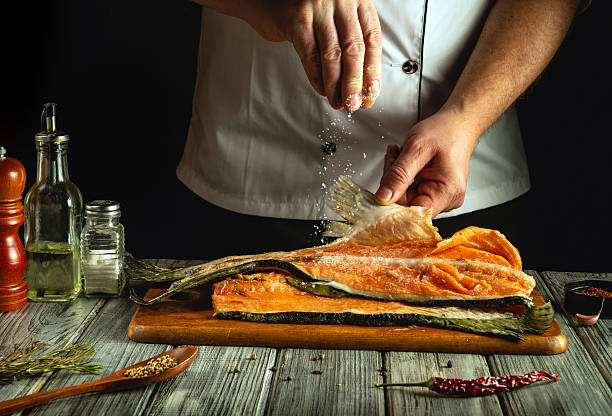 istock A chef skillfully prepares a large piece of fresh fish on a wooden cutting board, adding salt and spices to enhance the flavor. The kitchen is warmly lit with various cooking tools nearby 2219090767