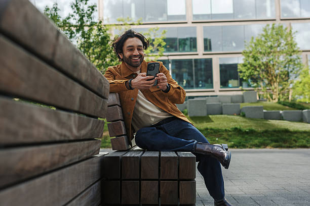 istock Smiling young man using smartphone while sitting on bench in city park 2219100500