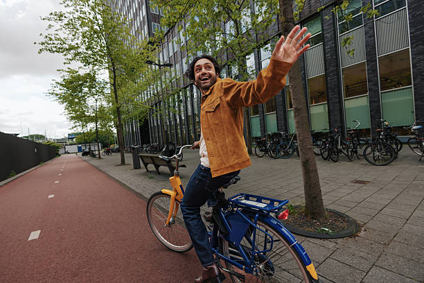 Young man joyfully waving while riding a bicycle along a scenic bike path in vibrant Amsterdam, Netherlands during a sunny day