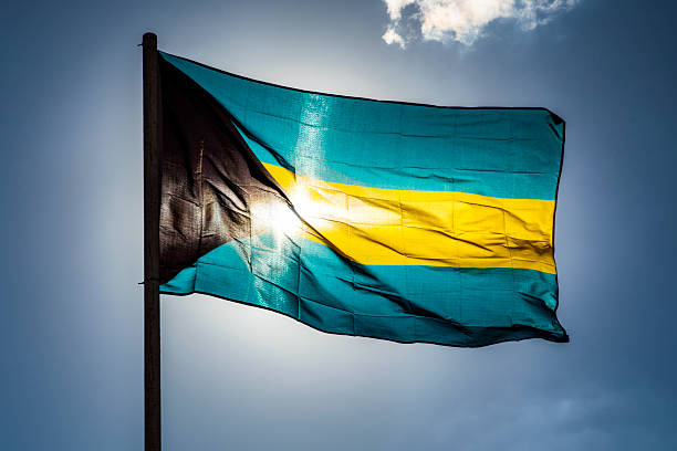Flag of the Bahamas with sun directly behind, wind blowing the flag straight out, mostly clear sky.