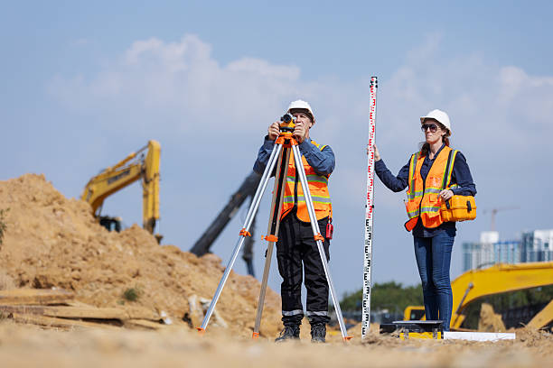 istock Two construction surveyors in safety gear stand at a job site using a tripod-mounted total station and leveling rod to measure land elevatio 2219116941