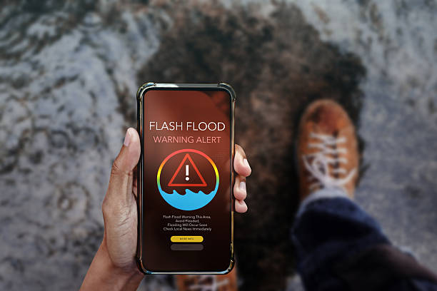 istock Smartphone displaying a flash flood warning alert screen, held by a person standing near rising floodwaters. Concept for emergency notification, natural disaster preparedness, and real-time weather alerts 2219124943
