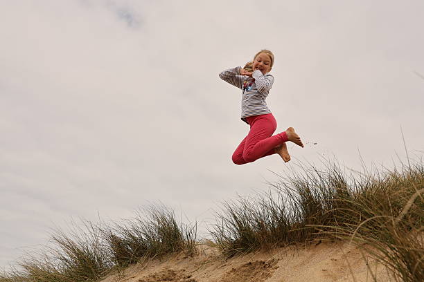 Young blonde haired girl jumping on marram grass covered sand dunes on a UK beach holiday