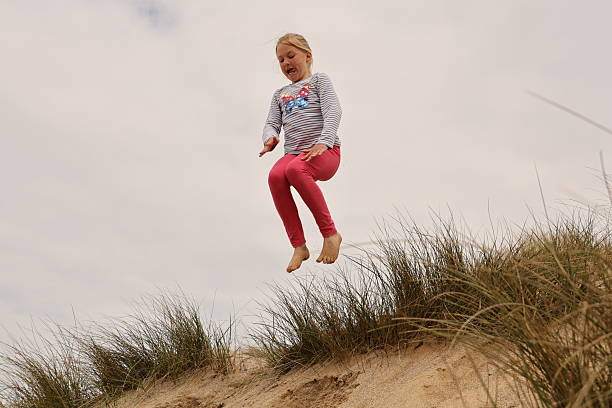 Young blonde haired girl jumping over sand dunes covered in Marram Grass on an overcast grey day