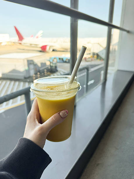 Stock photo showing close-up view of plastic, lidded, disposable cup, with a paper drinking straw, containing mango fruit juice smoothie being carried by an unrecognisable woman through an airport with a view of an aeroplane on the airport apron.