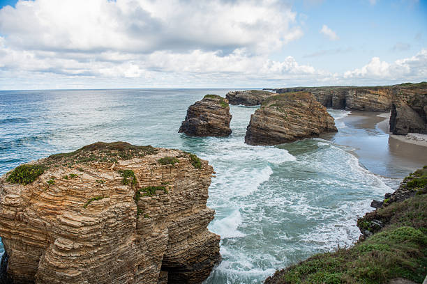 Coastal landscape showcases stunning rock formations and tranquil waves meeting the sandy shore, with a vibrant sky filled with clouds enhancing the natural beauty