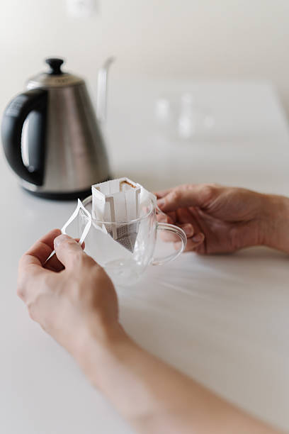 Vertical cropped shot of man carefully placing drip coffee bag into transparent glass cup, getting ready to brew fresh cup of coffee in modern kitchen setting at home