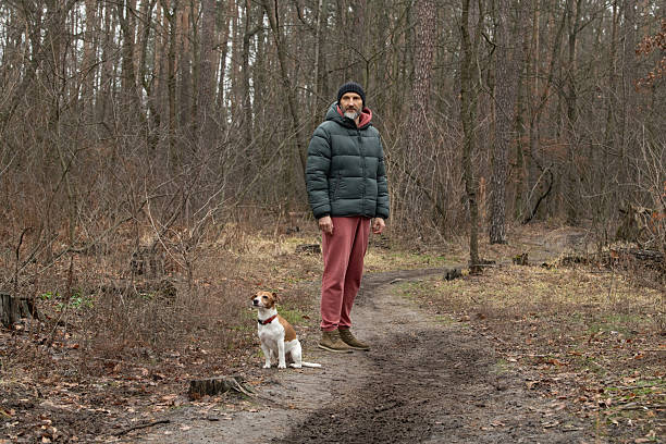 istock A man stands on a dirt path in a forest, dressed in a green puffer jacket and red sweatpants, accompanied by a small dog. The trees are bare, indicating late autumn 2219146938