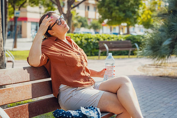 Displeased woman eyes closed sitting on a park bench holding a water bottle. Suffering from heat stroke and headache