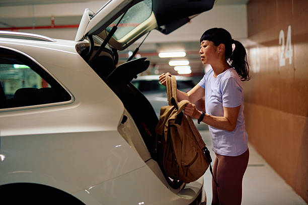 Asian mid-adult woman placing her backpack in the trunk of her white car after arriving at an underground parking garage, embodying an active lifestyle and convenience in urban commuting