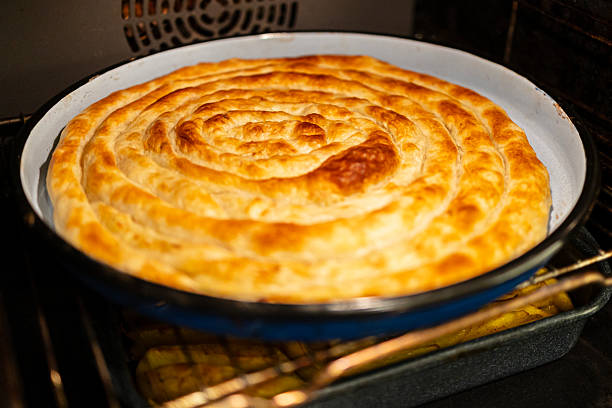 A close-up shot of a traditional Bosnian burek baking in a round metal tray inside the oven. The golden, flaky layers of pastry encase a rich filling of meat, cheese, or potatoes, creating a beloved savory pie enjoyed across Bosnia, Turkey, and the Balkans. The image captures the authentic process of preparation, showcasing the crisp texture forming as the burek bakes to perfection. Ideal for themes related to food promotion, traditional Balkan cuisine, bakery culture, homemade meals, and the growing popularity of comfort foods. A perfect visual representation of the irresistible appeal of local street food, rustic culinary traditions, and the enduring love for hearty, oven-baked delights.