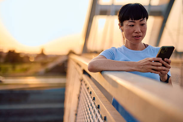 Asian sportswoman resting on a bridge at sunset, using her smartphone and enjoying music through wireless earphones after an intense training session. Embracing relaxation in the golden hour