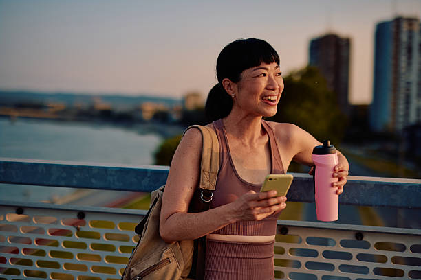 istock Smiling sportswoman using smartphone and holding water bottle on bridge at sunset 2219154939