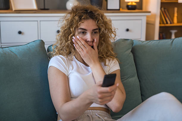 istock Young woman sitting on couch at home holding remote control, shocked and heartbroken while watching heartbreaking animal cruelty footage on television, emotionally affected by disturbing scenes 2219173403
