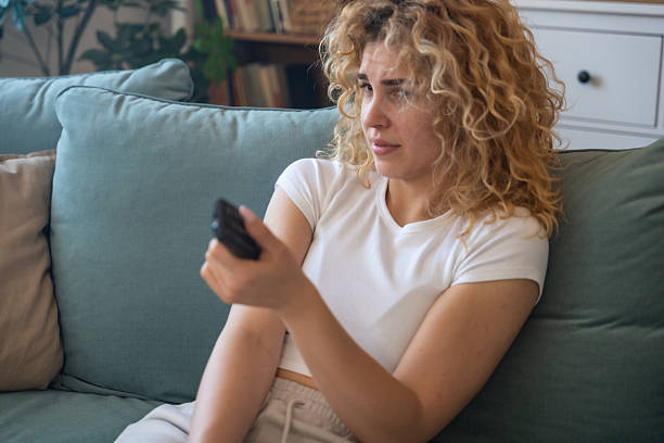 istock Young woman sitting on couch at home holding remote control, shocked and heartbroken while watching heartbreaking animal cruelty footage on television, emotionally affected by disturbing scenes 2219173404