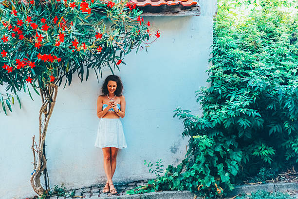 Tourist woman in a striped top and white skirt stands against a white wall, enjoying a peaceful summer moment under a tree with vibrant red flowers. She is using her smartphone for online dating, connecting with new people and exploring romantic possibilities through a dating app.