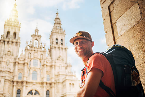 istock Joyful smiling middle-aged backpacker pilgrim sitting on Obradoiro square in Santiago de Compostela, Spain, with Santiago de Compostela Cathedral â famous Camino de Santiago finish point. 2219185183