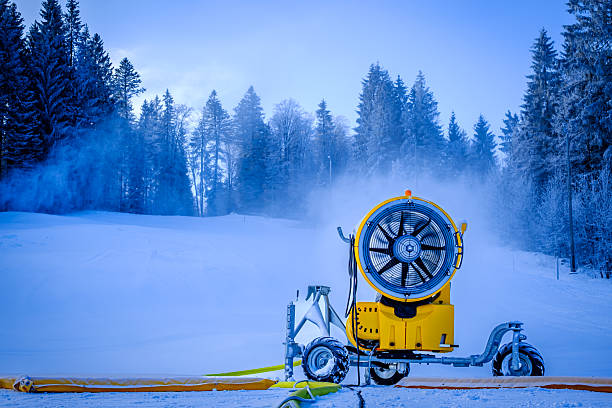 A snowmaking machine is actively producing artificial snow at a ski resort. The surrounding area features evergreen trees and a snowy landscape under a blue sky.
