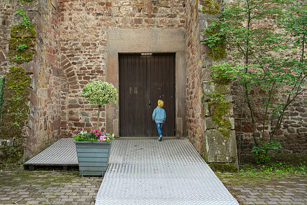 Young child wearing a yellow hat and blue jacket stands in front of a large wooden door set in a rustic stone wall, surrounded by greenery and potted plants on a cobblestone path.