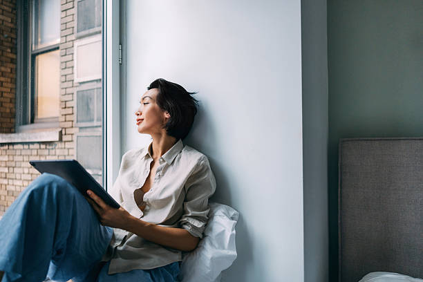 istock Woman Relaxing by Window Reading on Digital Tablet During Rainy Day 2219204204