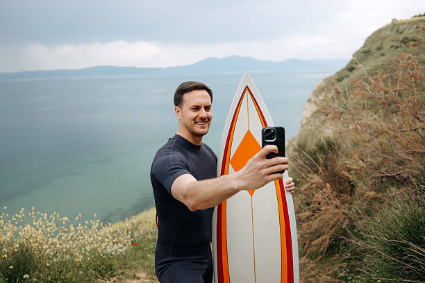 istock Smiling male surfer taking selfie while holding surfboard and standing on a cliff 2219205372