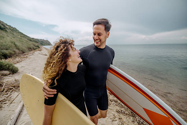 istock Smiling young couple in wetsuits walking at the beach while carrying surfboards 2219205436