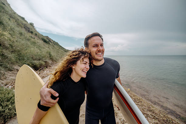 Smiling young couple in wetsuits walking at the beach while carrying surfboards