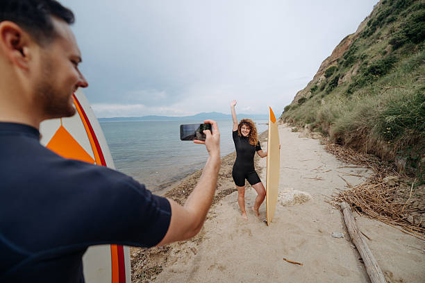 Man taking selfie of his girlfriend while she is standing on a beach with surfboard