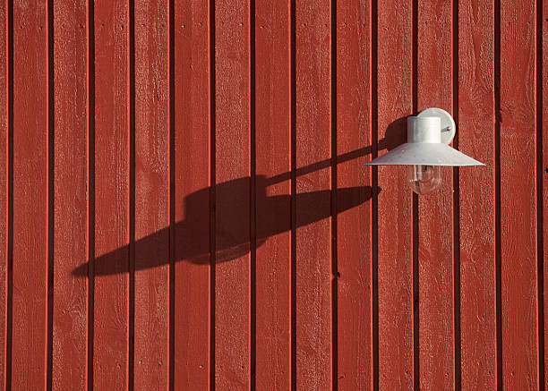 Sunlight highlights a white metallic lamp mounted on a vertical red wooden panel. The lamp casts a sharp shadow, emphasizing the texture and rustic charm of the wall's design.