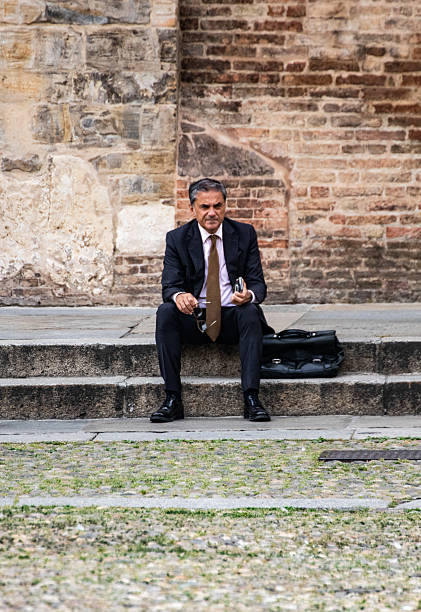 A businessman in a suit sits on a bench by a riverside in the evening holding a smartphone. The background features an urban setting with a focus on relaxation and contemplation