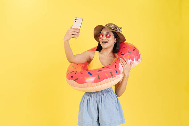 istock Cheerful young woman taking selfie with phone while wearing summer hat and donut float, dressed in casual vacation outfit, vibrant yellow background, fun travel or holiday concept photo 2219250209