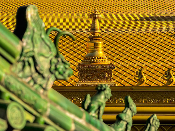 This image captures the intricate details of a traditional architectural roof, showcasing the craftsmanship and cultural heritage. The roof is adorned with a golden spire, which stands out against the textured yellow tiles that cover the roof's surface. The spire is embellished with elaborate patterns and designs that reflect a high level of artistic skill and attention to detail. In the foreground, there are green decorative elements with a dragon motif, adding a contrast in color and enhancing the overall aesthetic. The dragon is a symbol often associated with power, strength, and good fortune in many cultures. The warm lighting accentuates the rich colors and the metallic sheen of the gold, highlighting the textures and giving the scene a majestic and timeless quality. This photograph serves as a testament to the enduring beauty of traditional architecture and the importance of preserving such artistry for future generations.