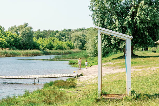 Public outdoor shower on grassy lake beach with people relaxing and playing near water on a sunny summer day.