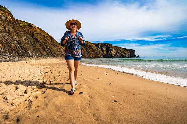 istock Middle aged woman in sun hat walking towards cliffs on sandy beach and enjoying holidays on spring day. Arrifana Praia, Portugal. Front view 2219259766