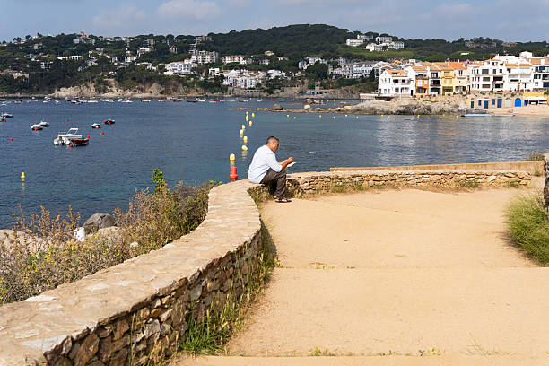Mature man sitting on stone wall, browsing smartphone along picturesque coastal landscape in Calella de Palafrugell, Catalonia, Spain