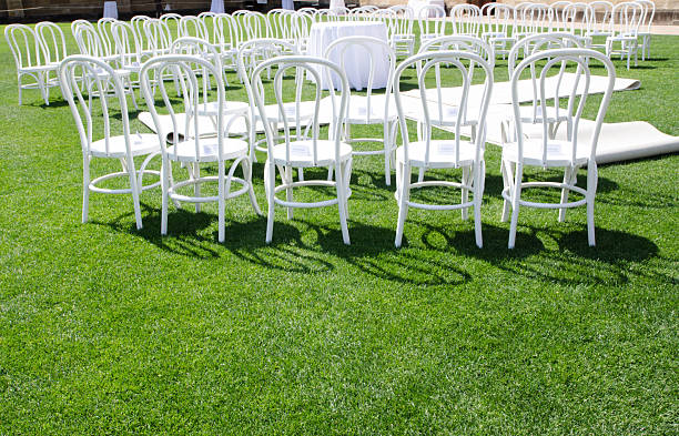 A group of White chair ceremony setting up on a green grass field, for a wedding or other formal event, a circular or semi-circular layout.