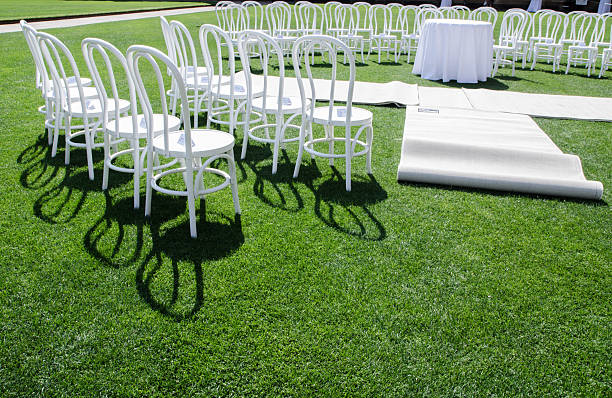 A group of White chair ceremony setting up on a green grass field, for a wedding or other formal event, a circular or semi-circular layout.