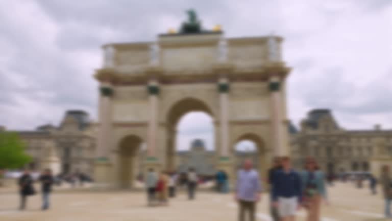 People stroll along the Place du Carrousel and take pictures in front of the Arc de Triomphe du Carrousel on a clear sunny spring day. Out of focus. Paris, France.