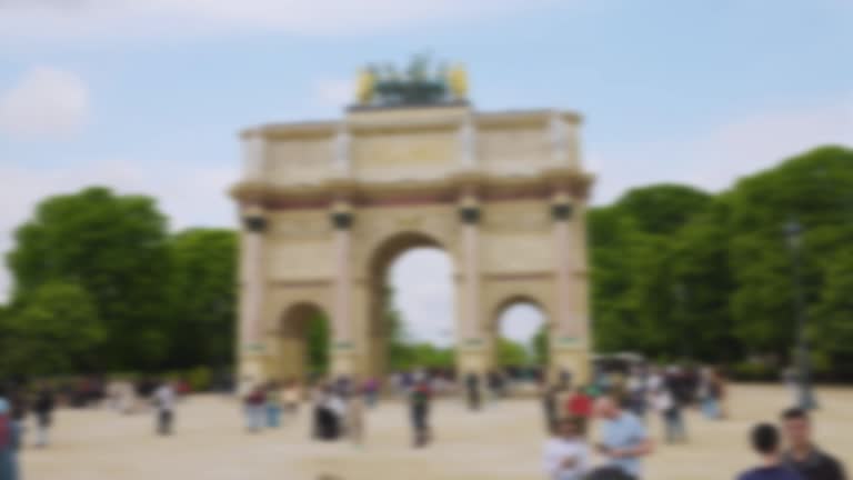 People stroll along the Place du Carrousel with a view of the Arc de Triomphe du Carrousel and the Jardin du Carrousel on a clear sunny spring day. Out of focus. Paris, France.