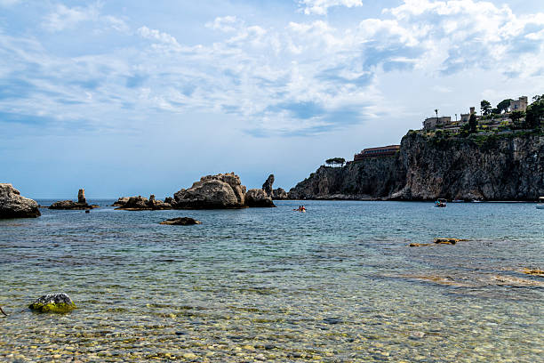 Taormina, Sizilien - Italy - 05-04-2025: Clear water with visible pebbles and rock formations near coastal cliffs and buildings, small boats floating on the calm sea near Taormina.