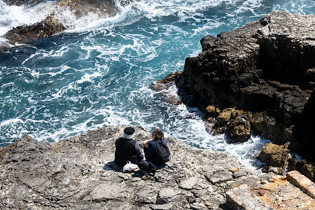 Syrakus, Sizilien - Italy - 04-09-2025: Two people sitting on rocky shore in Syracuse watching blue sea with white waves crashing against the coast under bright daylight.