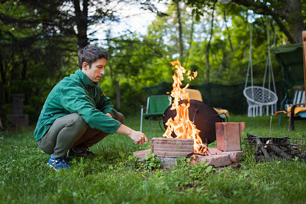 This photo captures a rugged lakeside cooking scene at golden hour. A campfire burns beneath a sturdy, blackened metal grill set atop a circle of large stones. The fire glows vividly with orange and red embers, while flames flicker upwards through the grates. Behind the setup, the sun is setting over a still lake, casting a bright flare and warm tones across the treetops. To the left, a black camping item—possibly a water bottle or pouch—rests on a rock, subtly suggesting human presence and the functionality of the space. The image communicates wilderness survival, warmth, and simple outdoor living.