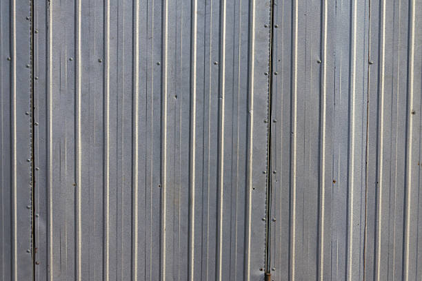 A close-up view of a corrugated iron wall, focusing on the repeating ridged pattern that defines its structure. The metal surface is lined with uniform, wavy grooves that create a rhythmic, undulating appearance across the wall. The material is industrial and durable, with a smooth yet rugged texture that reflects its functional design