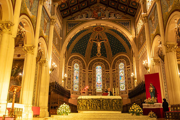 Reggio Calabria - Kalabrien - 05-22-2025: Man at ornate altar with crucifix and colorful stained glass windows, surrounded by columns, floral arrangements, and religious decorations.
