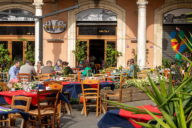 Catania, Sizilien - Italy - 04-15-2025: People sitting at colorful tables in an outdoor dining area in front of a pizzeria with decorative plants and flowers.