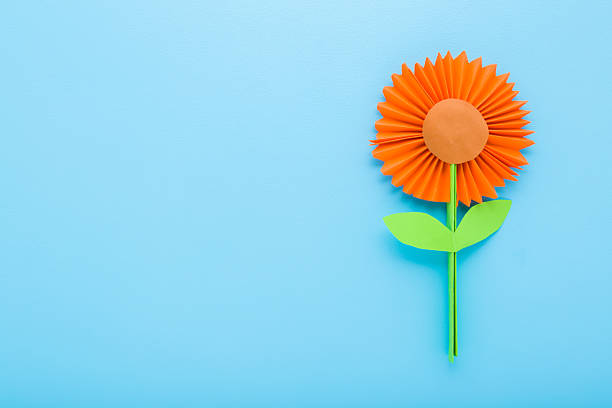 Orange flower with green leaves created from colorful application paper on light blue table background. Pastel color. Closeup. Empty place for text. Top down view.